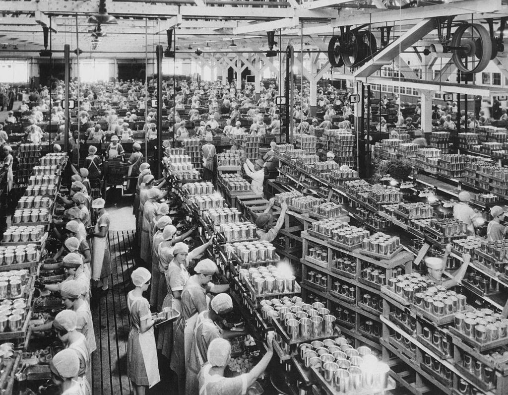 General view of a cannery in California, USA, circa 1940. The image shows rows of workers wearing hair nets alongside stacks of cans.