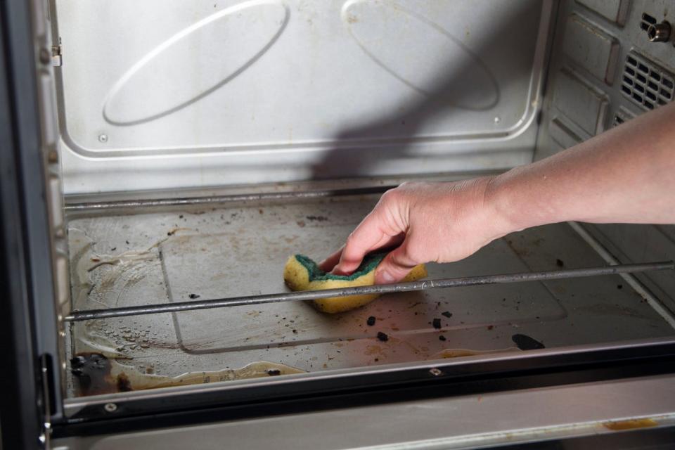 Woman's hand uses yellow and green sponge to clean oven grime before turning on self-cleaning function. 