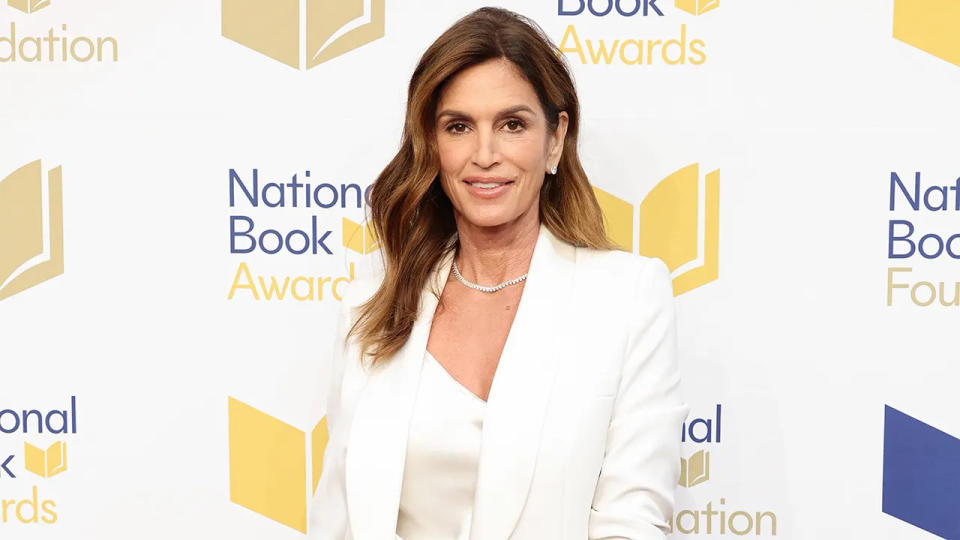 Cindy Crawford in a white tank top and white blazer smiles on the carpet