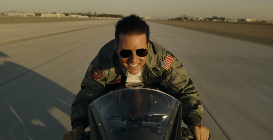 Tom Cruise is wearing sunglasses and a green jacket while riding a motorbike down an airstrip