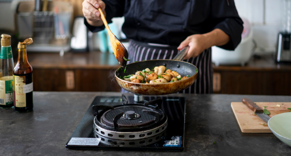 chef man in black shirt holding frying pan of potatoes over stove