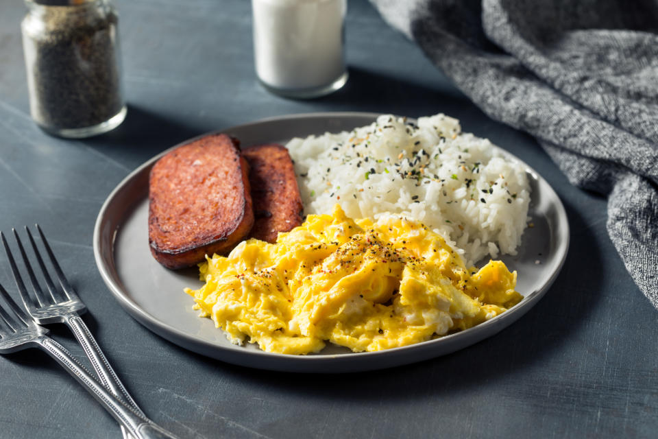 A plate of scrambled eggs, two slices of fried spam, and a serving of rice with sprinkled seasoning next to a fork, salt shaker, and a grey napkin