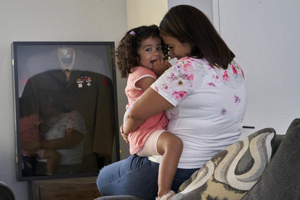 Laura Guerra and her daughter Emilia are reflected on her husband US Marine Lance Cpl. Rodrigo "Rigo" Guerra's uniform framed at their home in Riverside, Calif., on Monday, July 11, 2022. Rigo Guerra passed away on Dec. 24, 2020, due to complications following a monthlong battle with COVID-19. California has approved trust funds for some children from low-income families who lost a parent or caregiver to COVID-19. The Legislature set aside $100 million in the state budget to put into trust funds. (AP Photo/Damian Dovarganes)