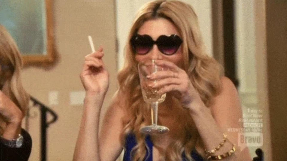 Brandi Glanville holding a cigarette and a glass of wine, wearing a sleeveless outfit and heart-shaped sunglasses