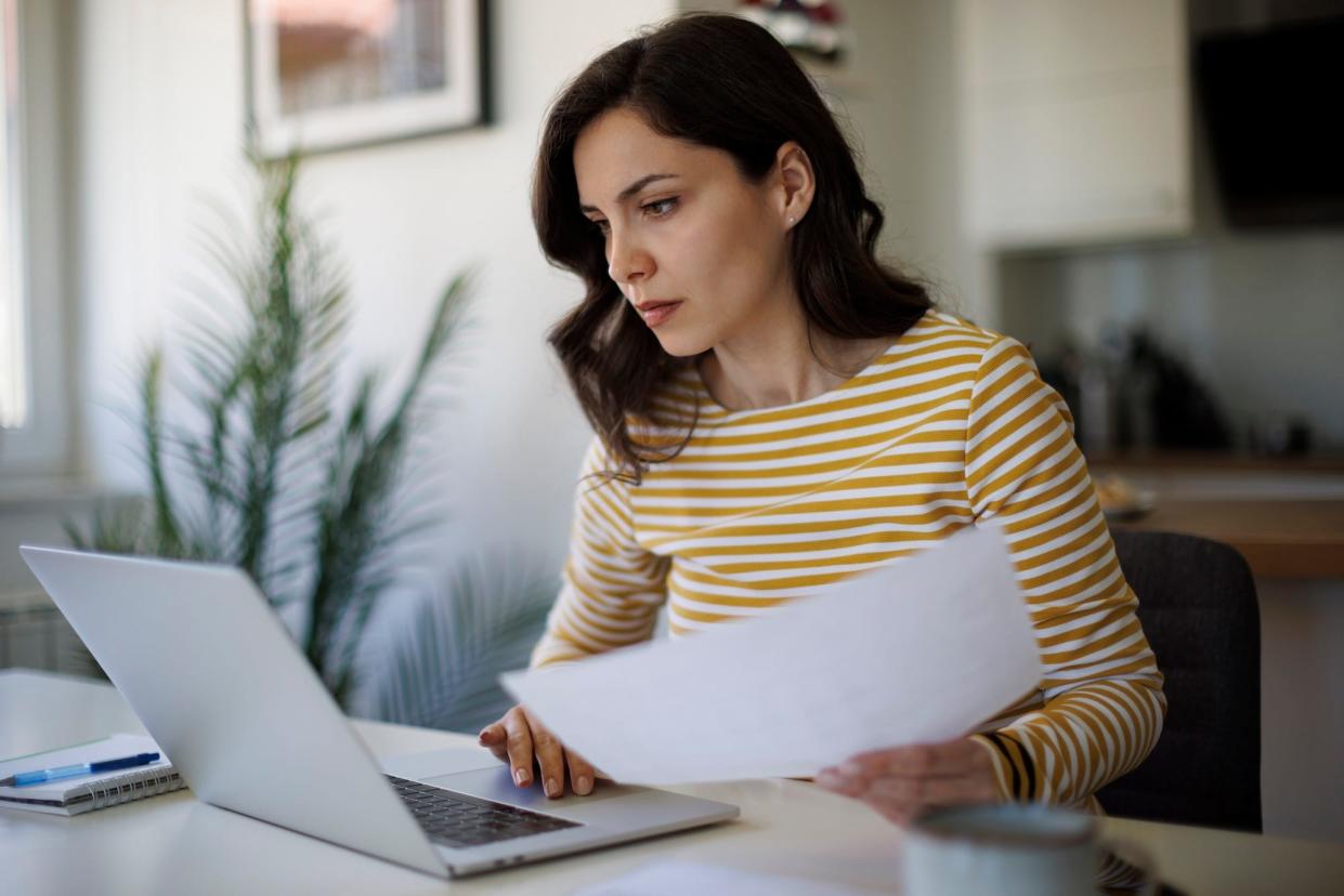 A woman working from home uses a laptop with one hand, and holds a piece of paper in the other.