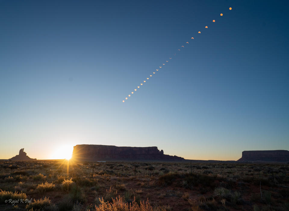 stages of the annular solar eclipse rise through the blue sky above large rock formations.