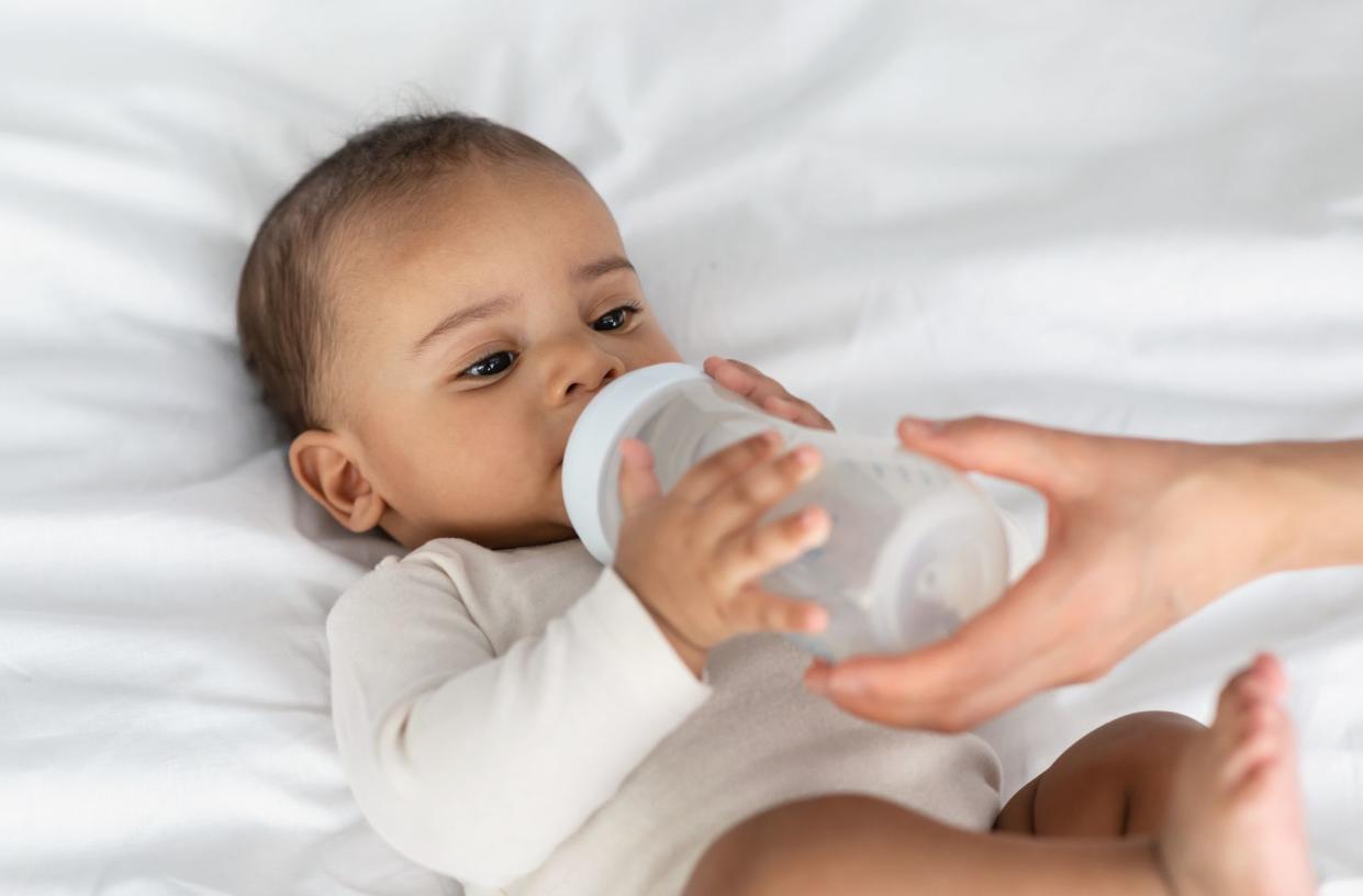 Childcare And Nutrition Concept. Portrait of cute baby drinking milk from bottle, woman helping and holding, kid wearing bodysuit lying on the white blanket at home