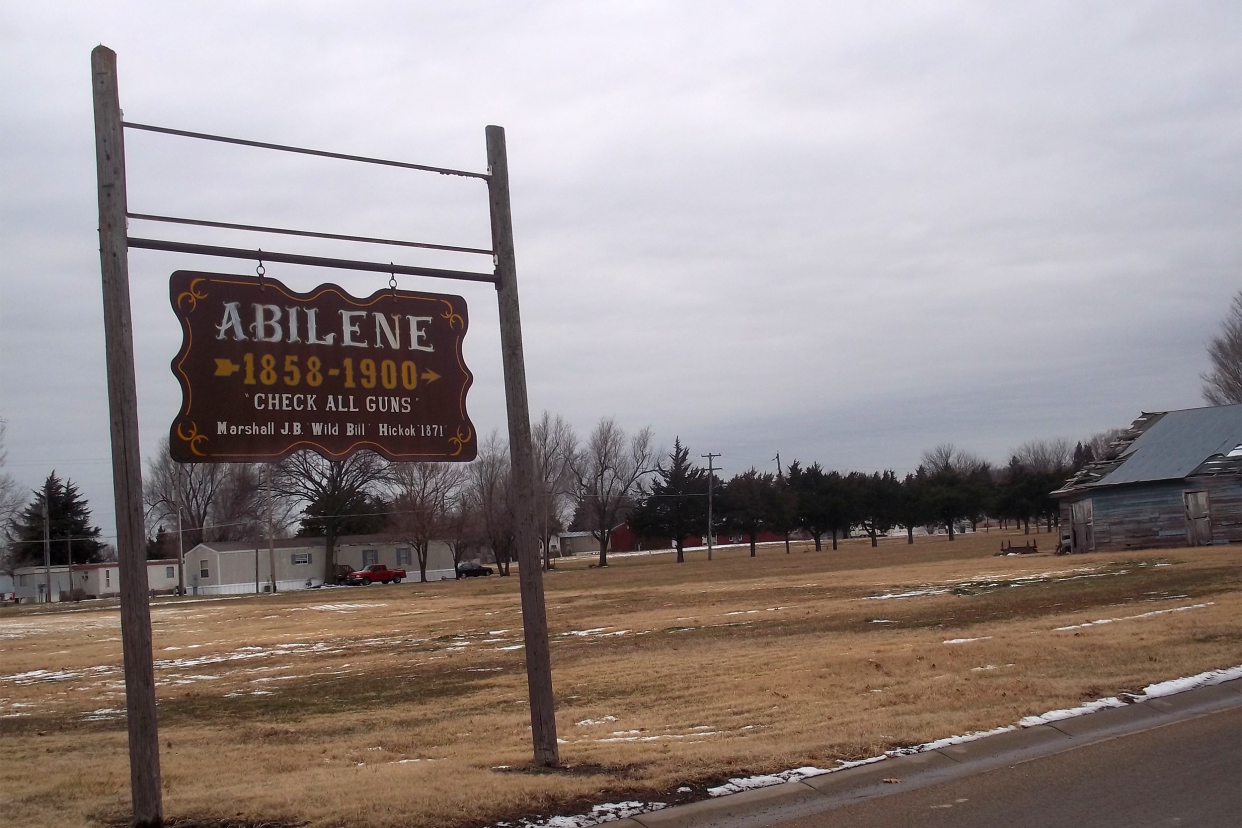 'Check All Guns' sign outside of the Historical Town of Abilene, Kansas, sign on the left, road on the right, lawn in the background, during winter, bare trees, grey sky, and slight snow on the ground