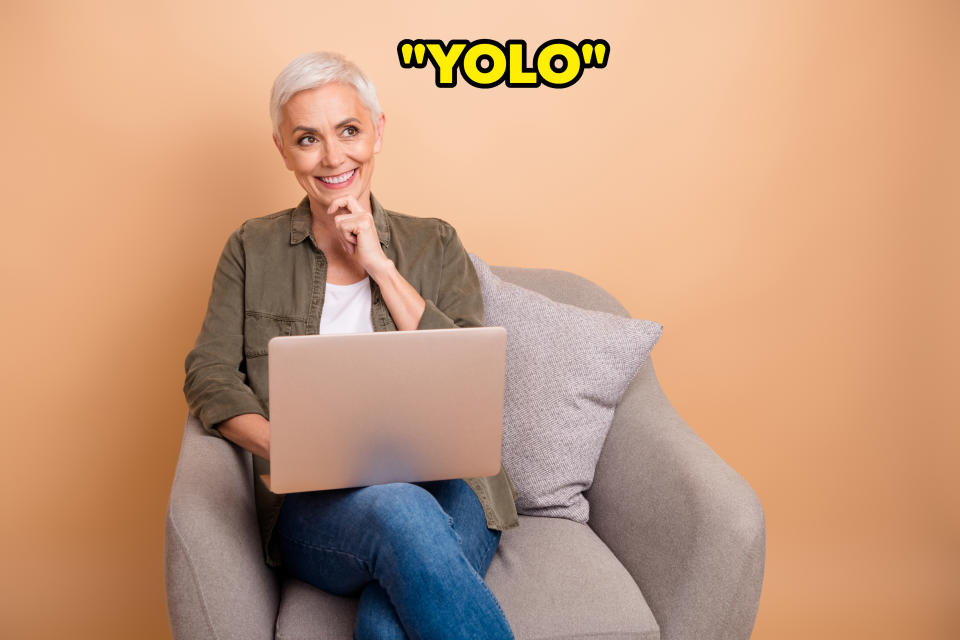 Woman with short hair seated with a laptop, smiling, resting chin on hand, wearing a casual jacket and jeans