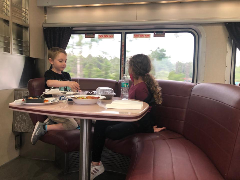 two kids sitting on burgundy booth on amtrak train sitting around a raised table with food on it