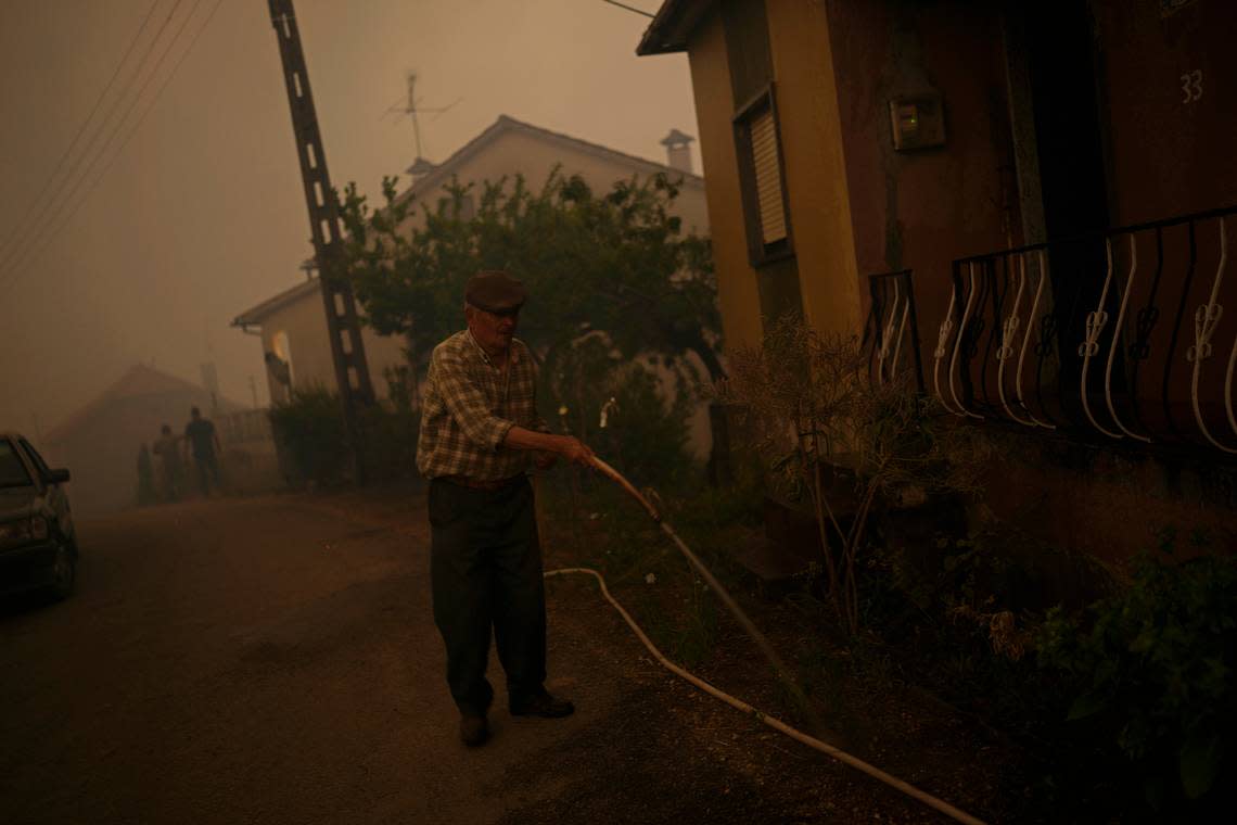 An elderly man uses a garden hose to water the outside of a house as a forest fire smoke darkens the sky in the village of Bemposta, near Ansiao, central Portugal, Wednesday, July 13, 2022. Thousands of firefighters in Portugal continue to battle fires all over the country that forced the evacuation of dozens of people from their homes. (AP Photo/Armando Franca)