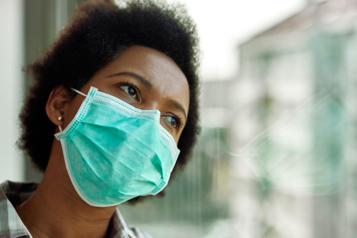 Distraught black woman with face mask thinking while looking through the window at home.