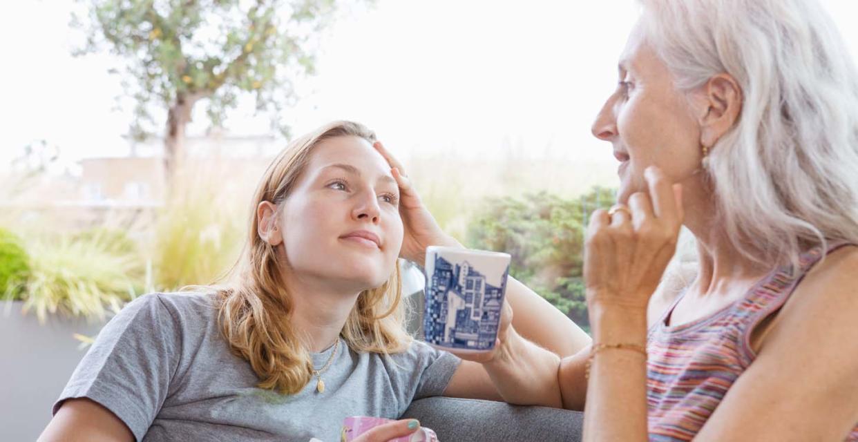 A mother and daughter sit facing each other, each holding a mug, with a tree and shrubs in the background.