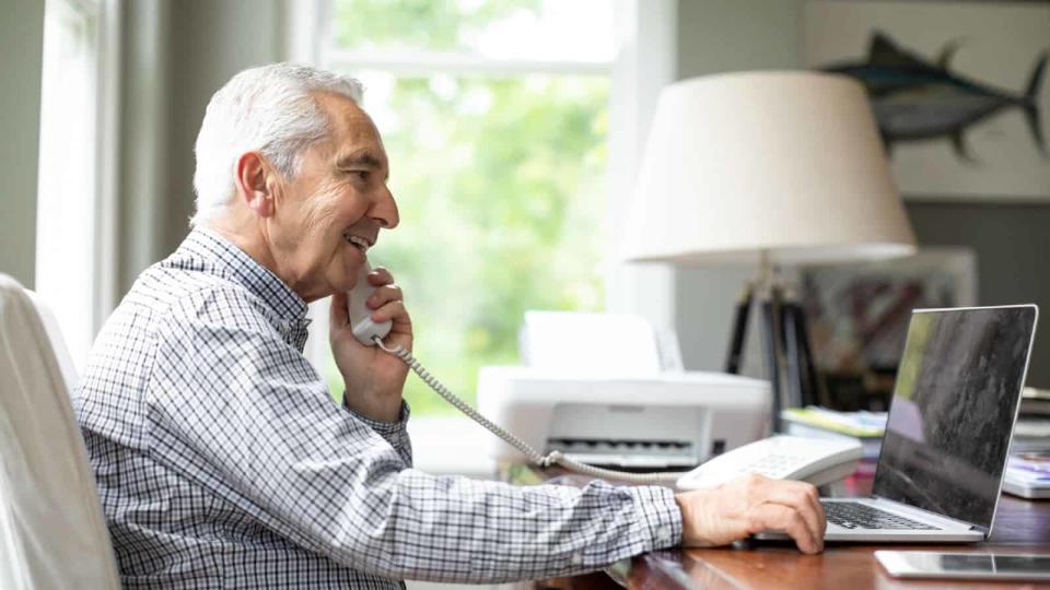 Smiling senior white man talking by phone while using laptop in office.