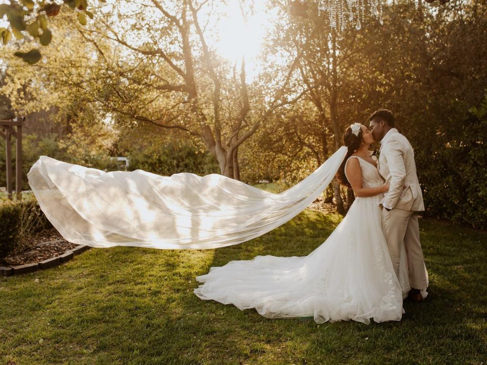 A bride and groom kiss in a field as the bride's veil flies up.