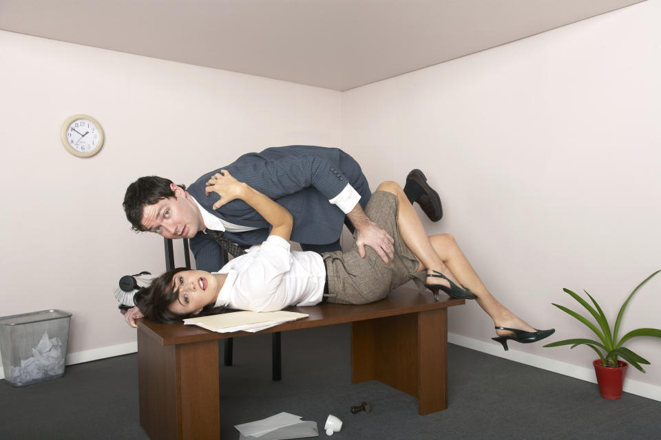 Two people are sprawled on a desk in an office, one in a suit and the other in a skirt and blouse, with some papers and a pot plant nearby