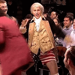 A person is dressed in vintage attire with an 18th-century coat, vest, and American flag shorts, dancing on SNL