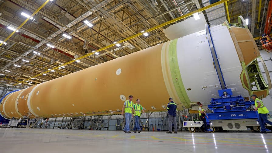  orange rocket stage lies on its side in a big hangar. white stripes mark the edge of the rocket and a machine of some sort sits in front of it. technicians walk in front of the rocket with yellow safety vests 