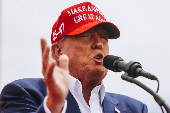 Donald Trump speaks into a microphone and wears a "Make America Great Again" Hat and a suit jacket