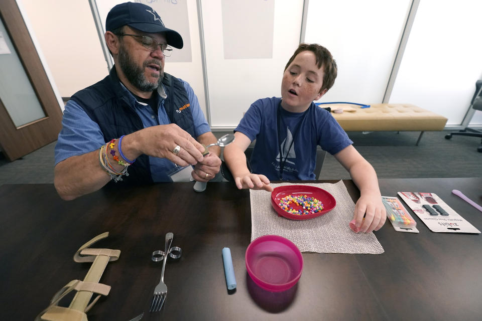 Charlie Warlick, right, gets an explanation from Tom Simon on using modified eating utensils during a workshop for young caregivers of ALS diagnosed family members in Dallas, Texas, Saturday, April 9, 2022. The children have gathered for a clinic to learn more about caring for people with Lou Gehrig's disease, or amyotrophic lateral sclerosis. It's a fatal illness that attacks nerve cells that control muscles throughout the body. (AP Photo/LM Otero)