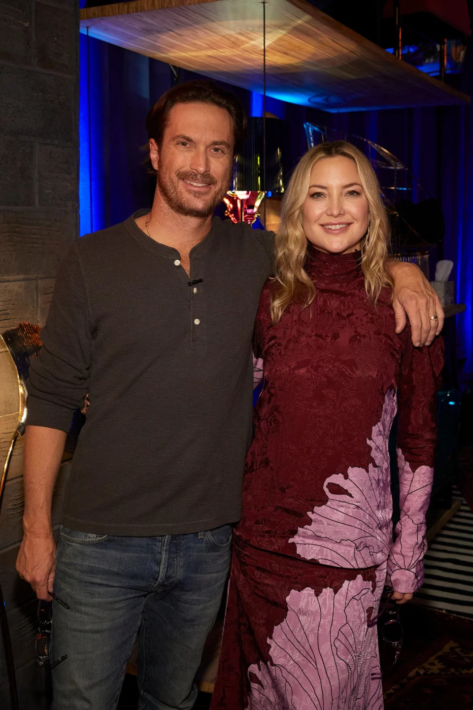 Oliver Hudson and Kate Hudson pose together, with Oliver in a casual shirt and jeans, and Kate in a long dress with floral patterns