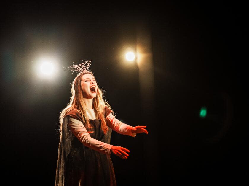 A woman illuminated by stage lights opens her mouth wide as she acts. 