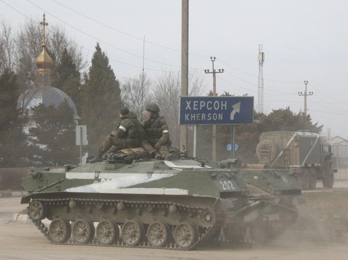 Two servicemen ride on a Russian armored vehicle marked with the letter Z, passing a sign pointing to Kherson.