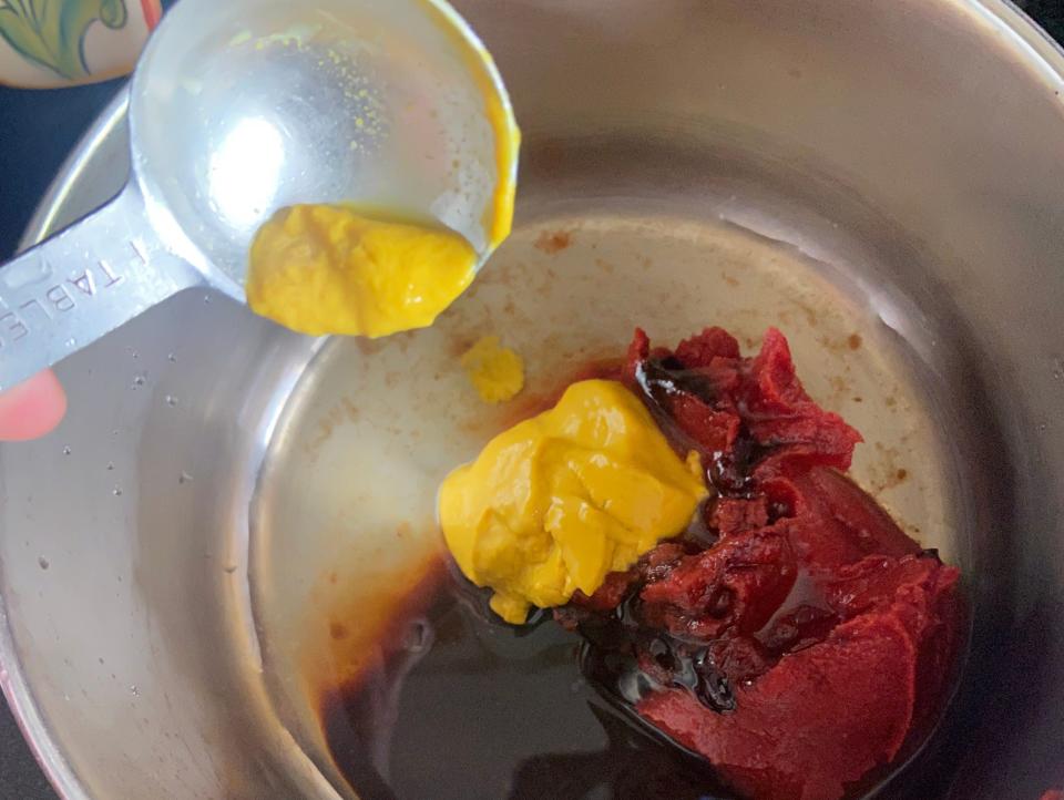 hand pouring yellow mustard into a pan with ingredients for barbecue sauce