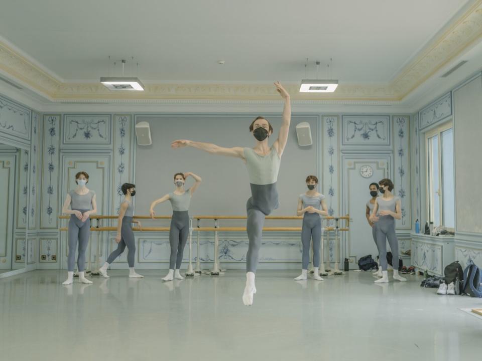 A ballet student takes a leap while other students watch and warm up in the background