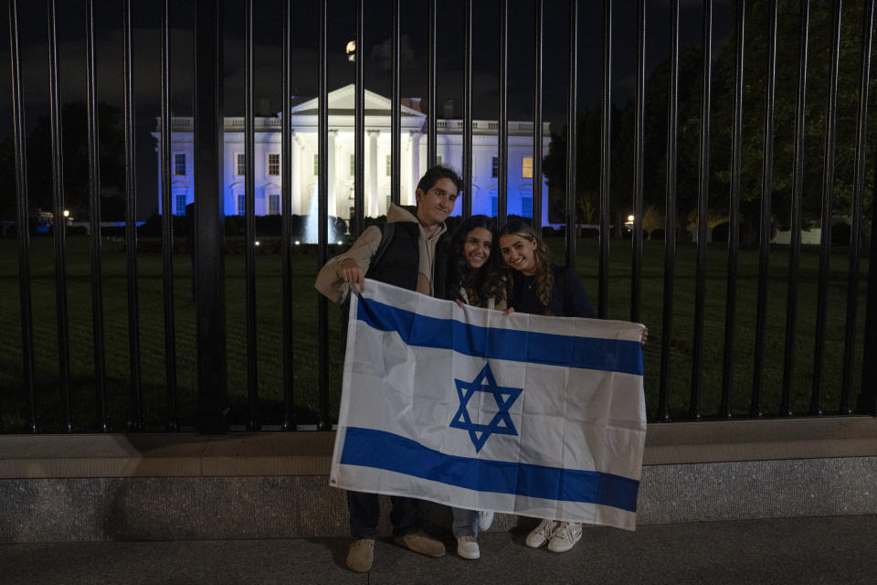 People stand with a Israeli flag outside the White House, lit in blue and white to underscore U.S. solidarity with Israel, Monday, Oct. 9, 2023, in Washington. (AP Photo/Jon Elswick)