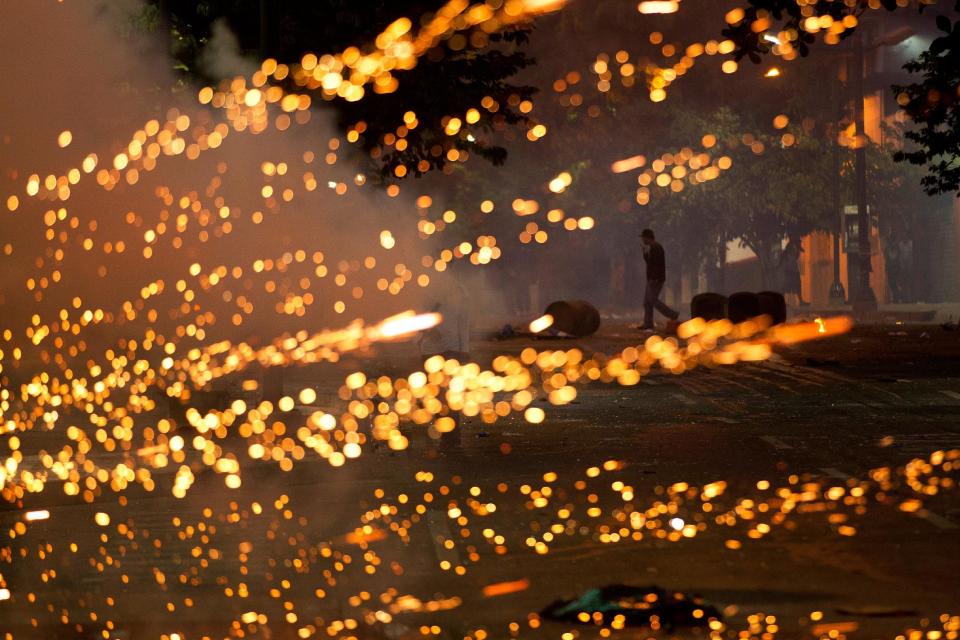 An anti-government protester walks near a burst of lights caused by a fire cracker launched by protesters at Bolivarian National Policemen during clashes in Caracas, Venezuela, Saturday, March 22, 2014. Two more people were reported dead in Venezuela as a result of anti-government protests even as supporters and opponents of President Nicolas Maduro took to the streets on Saturday in new shows of force. (AP Photo/Esteban Felix)