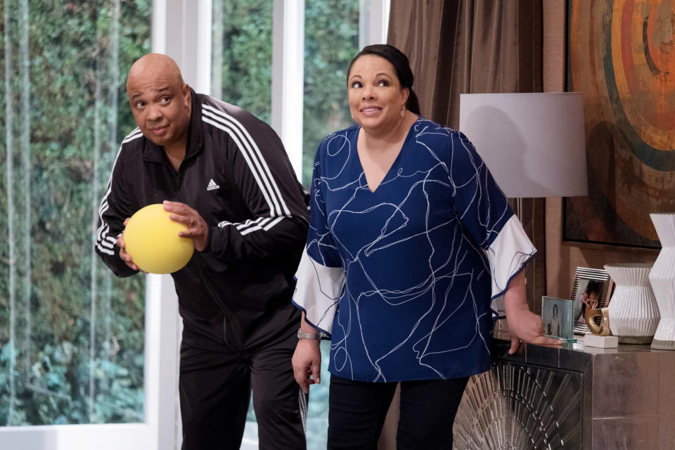 Rev Run, in a tracksuit, prepares to throw a yellow ball while Justine Simmons, in a patterned blouse, stands beside him inside a modern living room