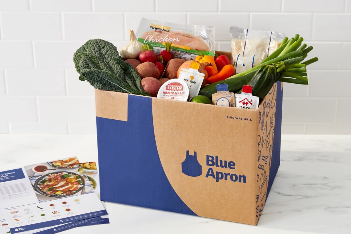 A blue apron box sits on a kitchen counter with fresh produce in it such as kale, tomatoes, green onions, etc. 