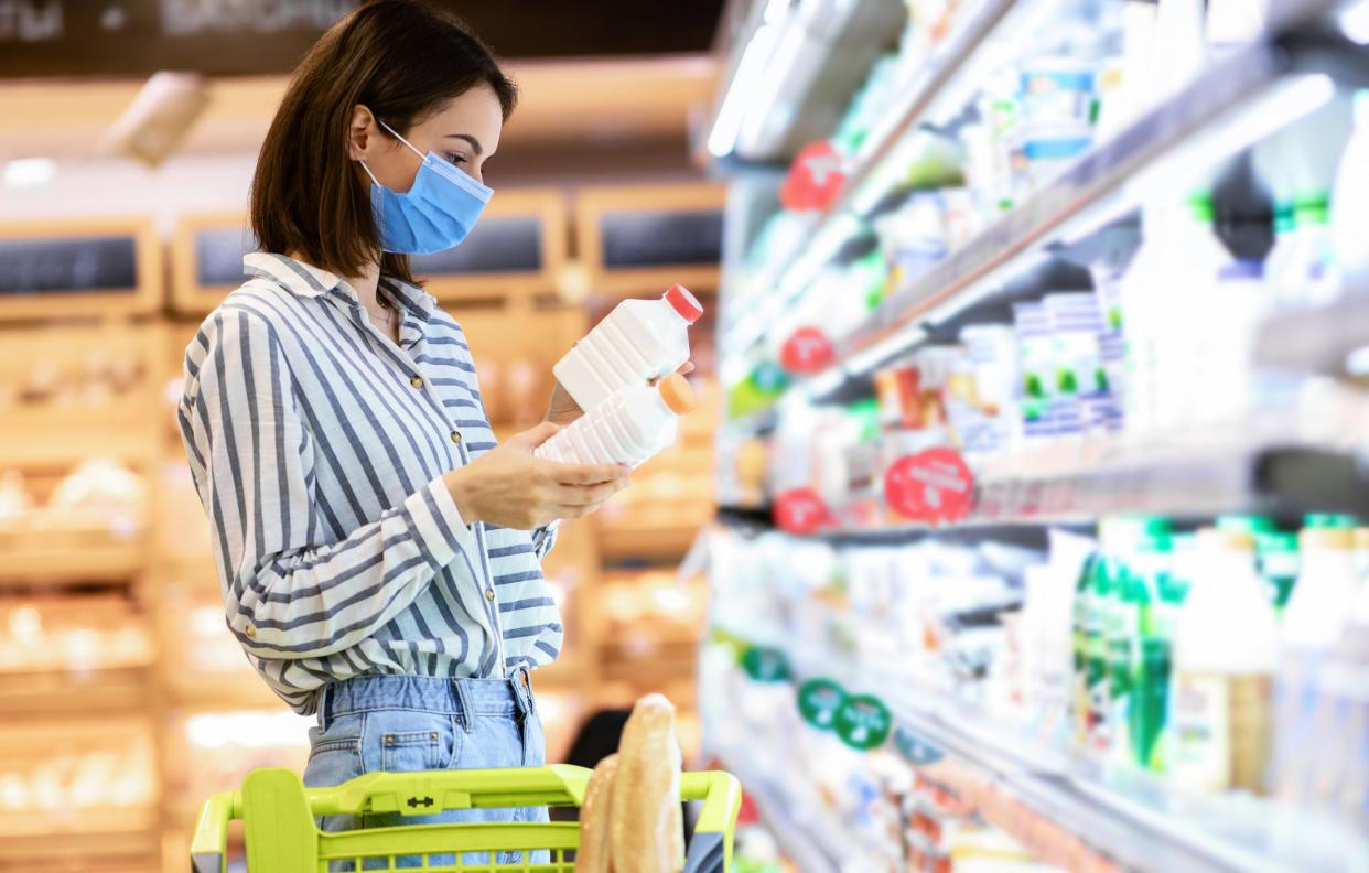 Consumerism. Young woman in medical disposable mask holding two bottles of milk or yoghurt, standing near the freezer, choosing dairy products, making decisions, checking expiry date