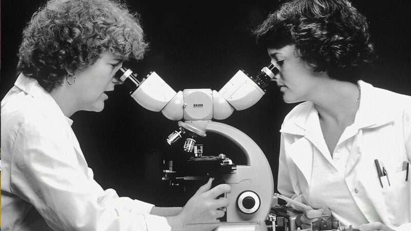 A black and white photo of two people in white coats looking into microscopes