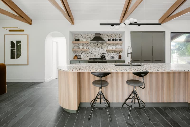 Wide angle of a contemporary modern kitchen with terazzo tiles and countertop, fluted wood island, industrial-style barstools, with gray floors, white wood ceilings, and natural wood rafters.