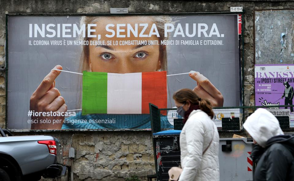 People walk past a huge billboard that shows a woman with as protective mask in the colours of an Italian flag and which reads, "All together, without fear" referring to the coronavirus campain to stay home in the city of Naples on March 22, 2020. - Italian Prime Minister Giuseppe Conte on March 21, 2020, ordered all non-essential companies and factories to close nationwide to stem a coronavirus pandemic that has killed 4,825 people in the country in a month. "The decision taken by the government is to close down all productive activity throughout the territory that is not strictly necessary, crucial, indispensable, to guarantee us essential goods and services," Conte said in a late-night TV address (Photo by Carlo Hermann / AFP) (Photo by CARLO HERMANN/AFP via Getty Images)