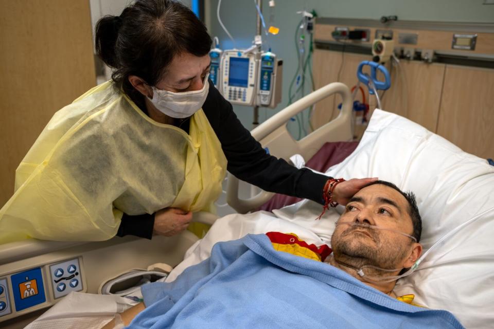 A masked woman touching the head of her brother as he lies intubated on a hospital bed