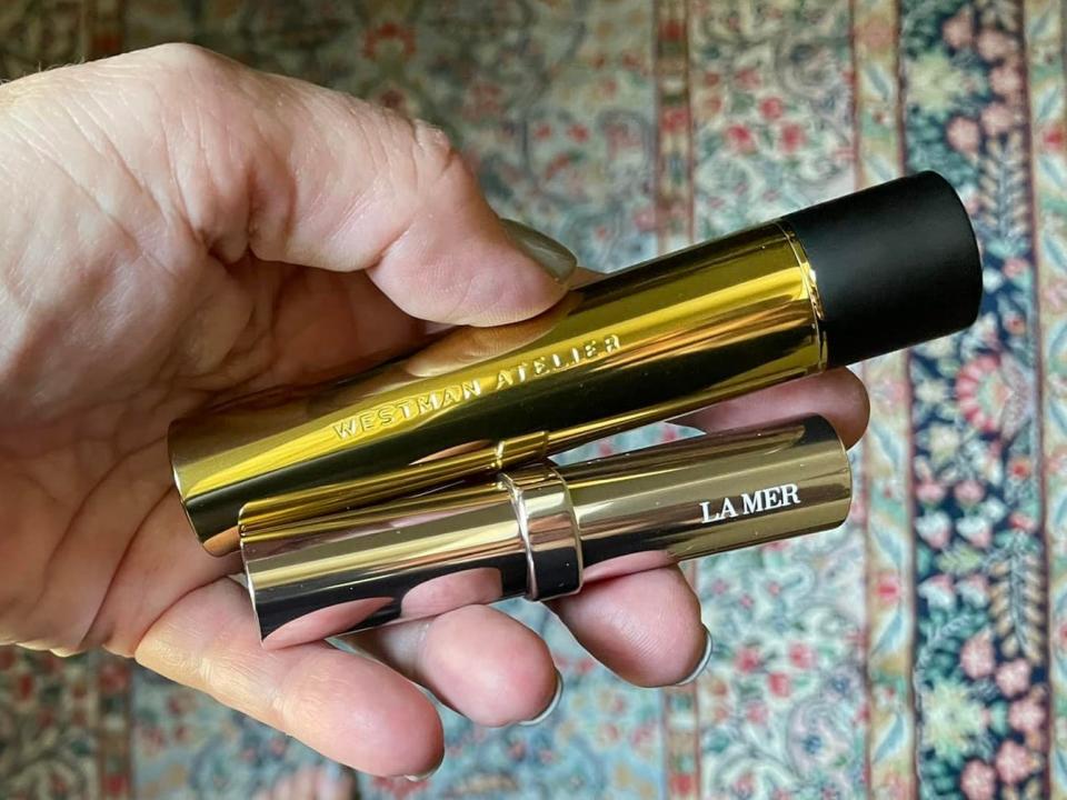 The writer holds a golden tube of mascara and a tube of stick concealer with "La Mer" on the side of it