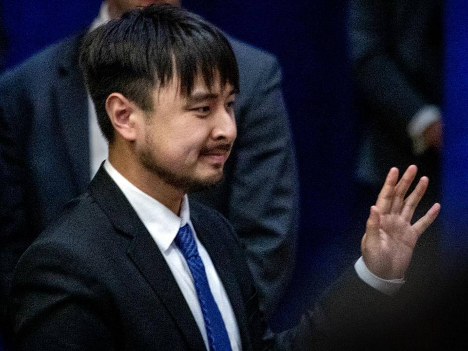 A man with dark hair, in a dark suit and blue tie, waves