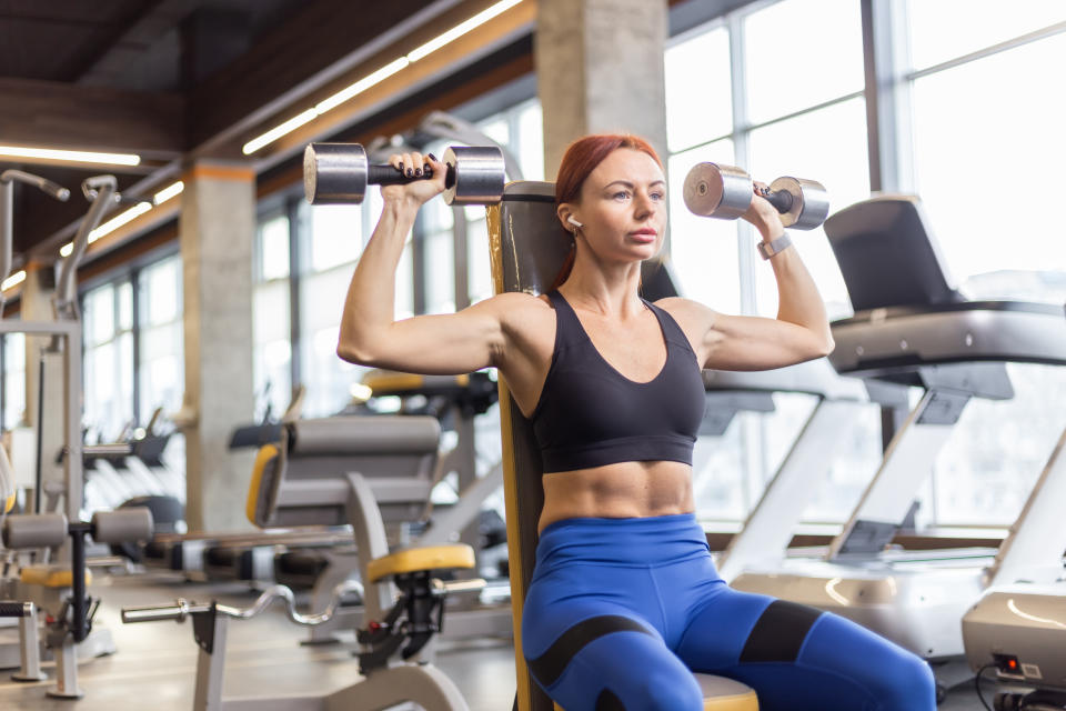 Fit woman trains shoulders with dumbbells in her hands doing dumbbell bench press over her head while sitting on a bench in the gym