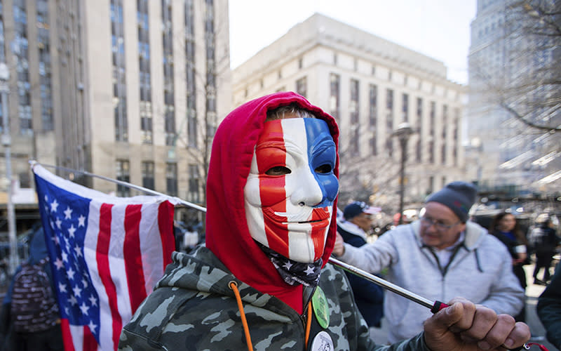 A man wearing a patriotic colored Guy Fawkes mask and holding an American flag, joins a small group of protesters near the Manhattan District Attorney's office