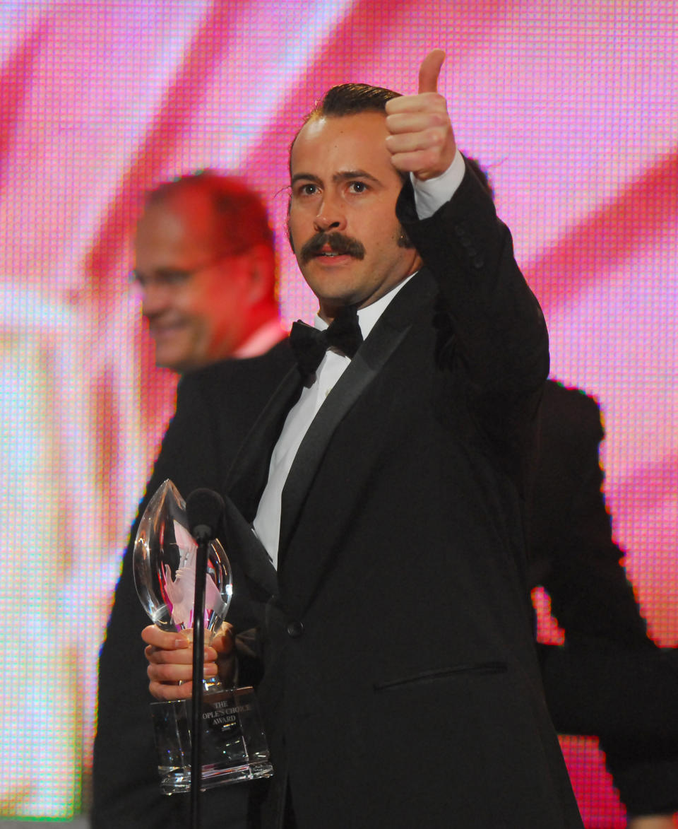 Jason Lee in a tuxedo gives a thumbs-up and holds a trophy at an awards event