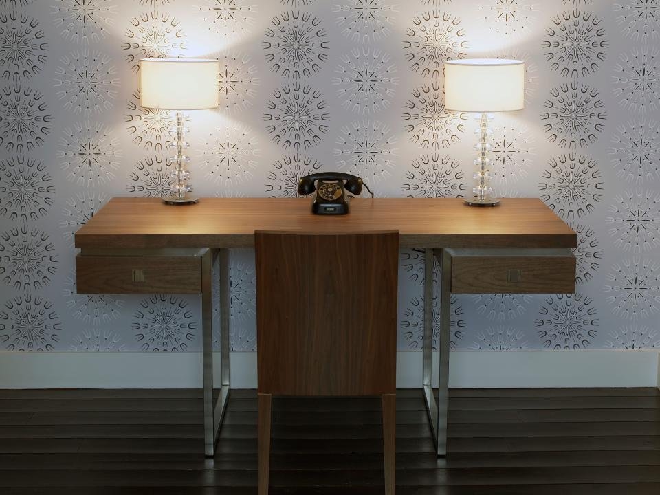 Wooden desk and chair with telephone centered on top and two matching lamps in front of lavender and brown patterned wallpaper