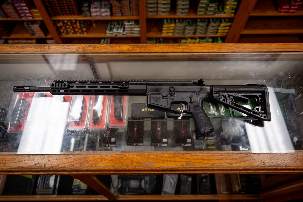 A long black gun is displayed in a case. Shelves full of gun equipment surround the case. 