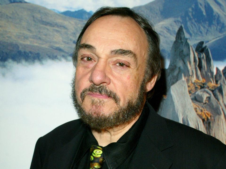 John Rhys-Davies wearing a black button down and standing in front of a fantastical background