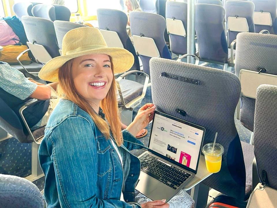 Photo of the writer opening a laptop and sitting on a seat on a ferry. She wears a straw hat and a blue jeans jacket.