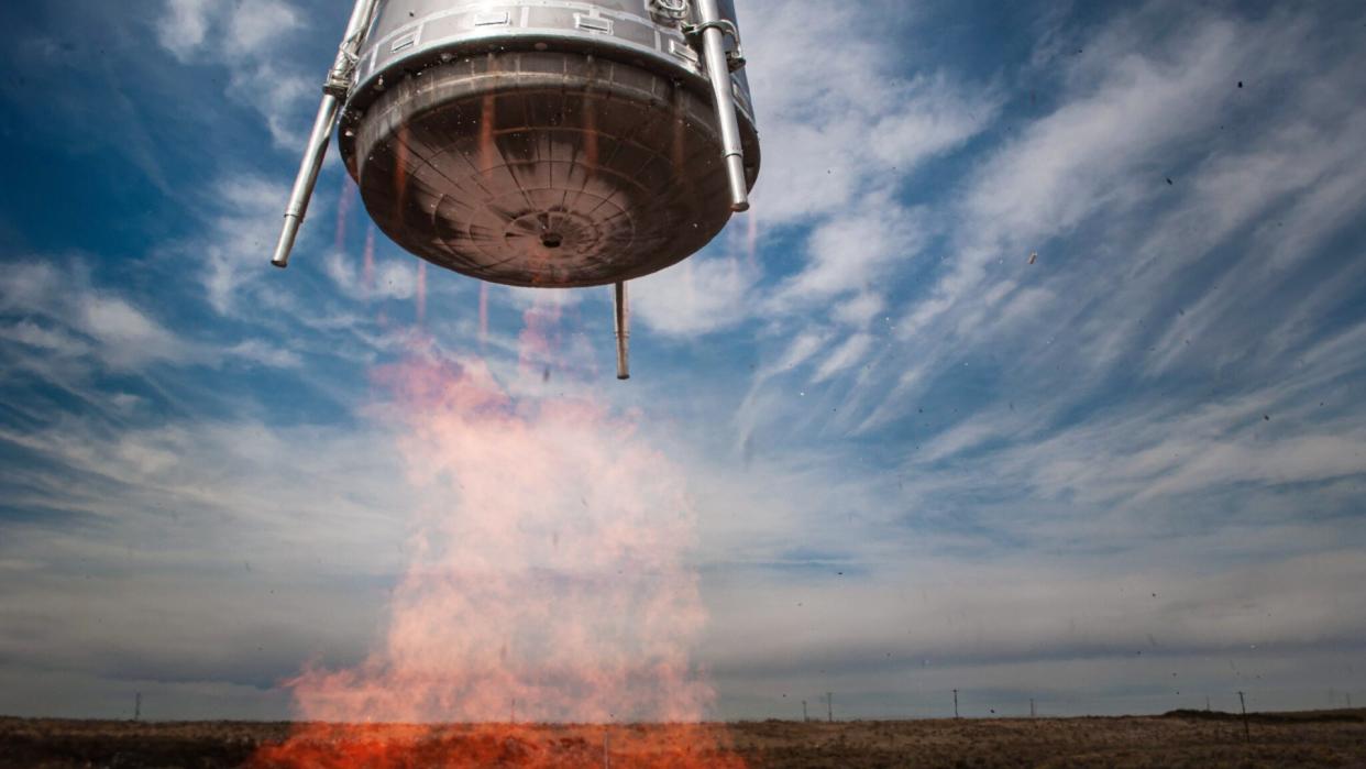  A squat metal cylinder hovers above the ground with a plume of fire beneath it. 