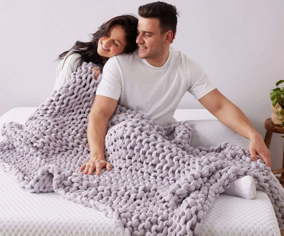 A man and woman sit and snuggle under a mauve calming blanket and a matress.