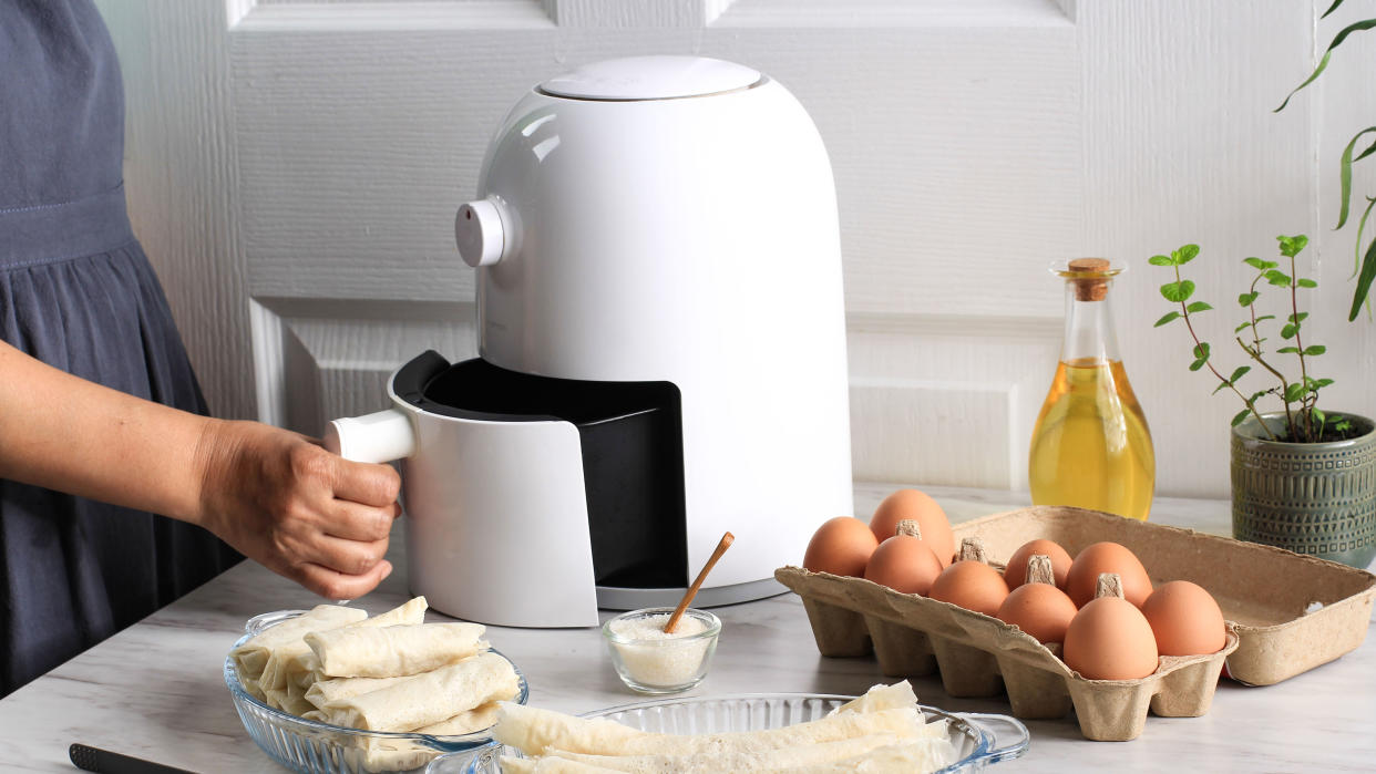  A white air fryer with the basket opened by hand on a table with surrounding ingredients, including eggs and oil . 